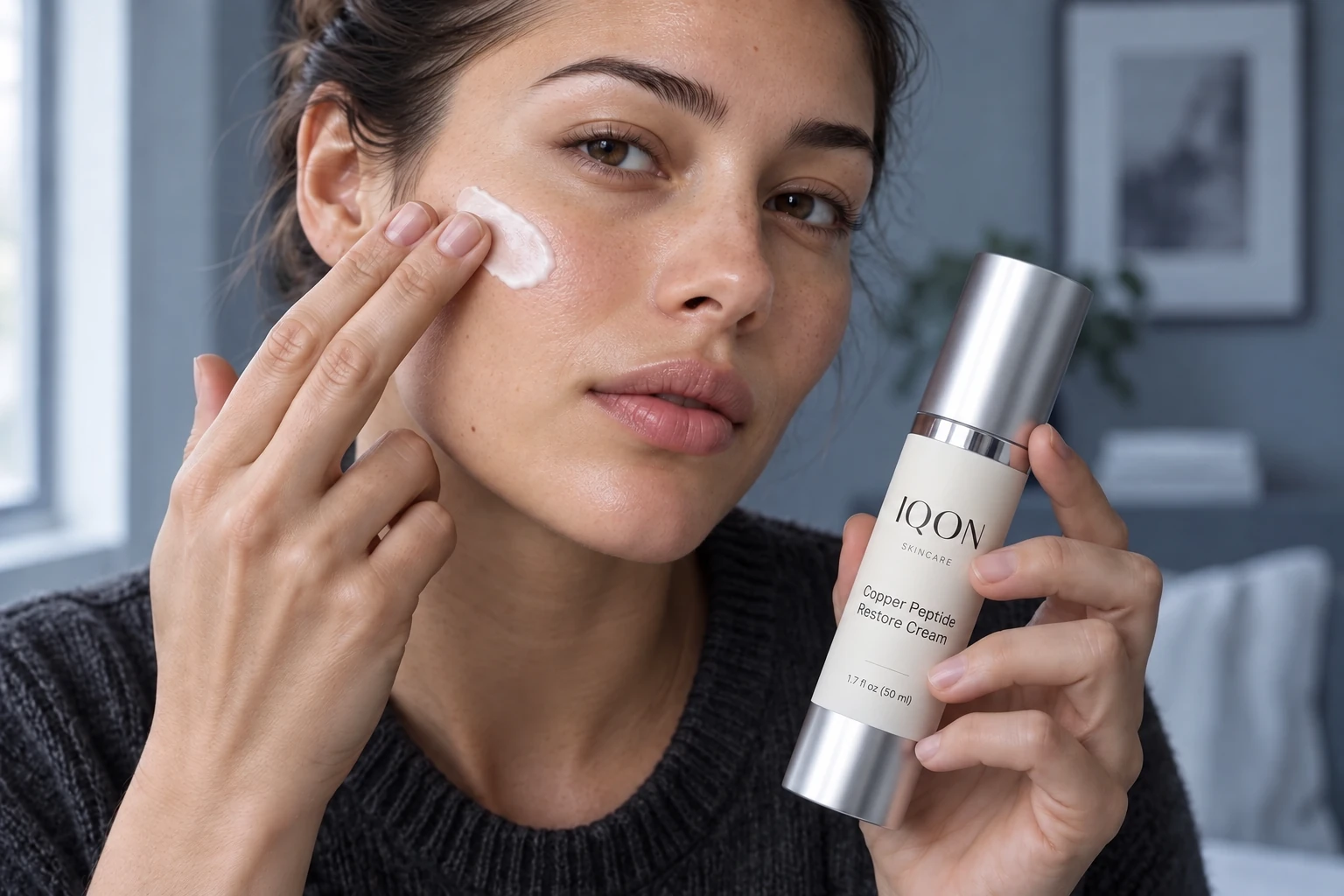 A woman applying cream to her cheek while holding IQON Copper Peptide Restore Cream